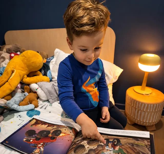 A boy in bed pointing at an illustration in his personalized book by lamplight